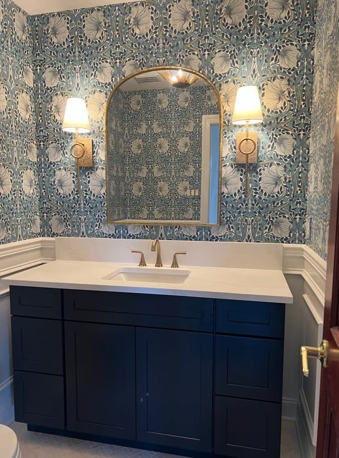 Powder room with navy vanity, patterned wallpaper, brass sconces, and arched mirror.