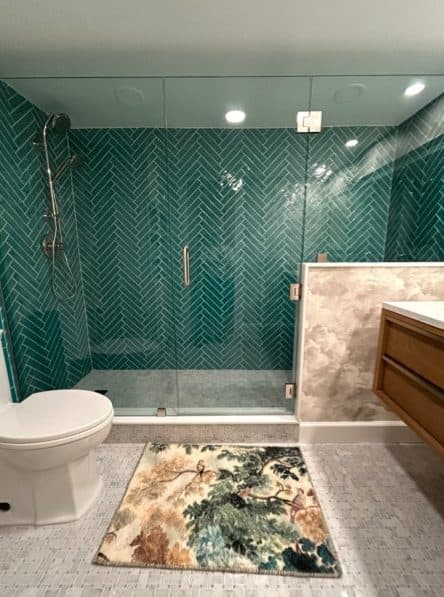 Bathroom shower with teal tile, glass enclosure, and patterned floor runner.